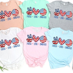 May include: Five t-shirts in different colors with the same design. The design features a peace sign, a heart, and a circle with the American flag pattern. The text "Peace Love America" is printed below the design.
