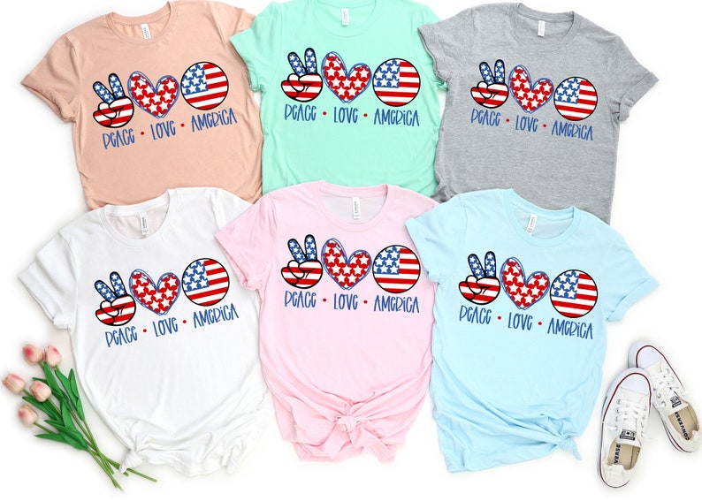 May include: Five t-shirts in different colors with the same design. The design features a peace sign, a heart, and a circle with the American flag pattern. The text "Peace Love America" is printed below the design.