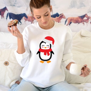 May include: White sweatshirt with a cartoon penguin wearing a red Santa hat and scarf. The penguin is smiling and has black eyes.