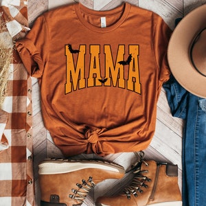 May include: Orange t-shirt with the word "MAMA" in orange block letters with black outlines and bat graphics. The shirt is styled with a brown hat, jeans, and boots, and a plaid shirt.
