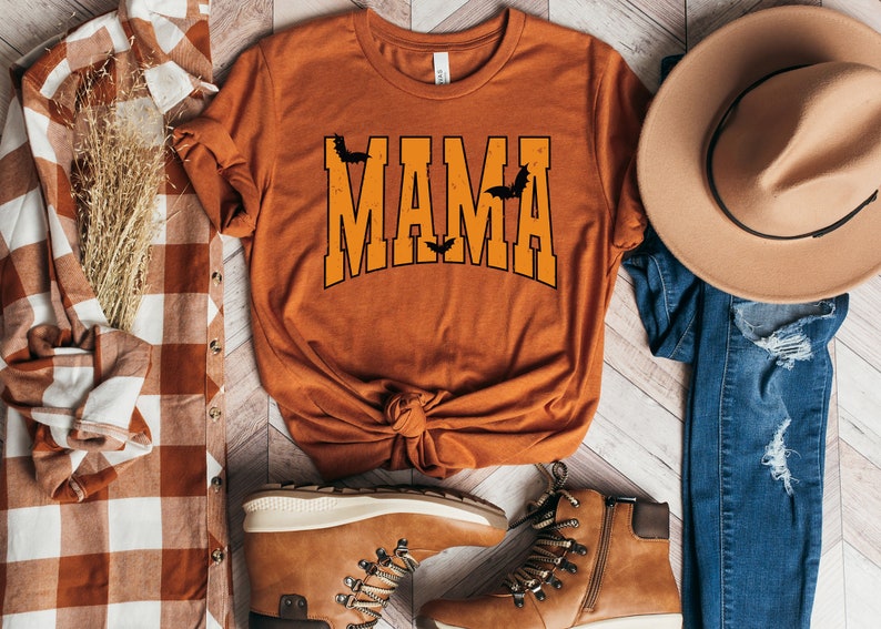 May include: Orange t-shirt with the word "MAMA" in orange block letters with black outlines and bat graphics. The shirt is styled with a brown hat, jeans, and boots, and a plaid shirt.