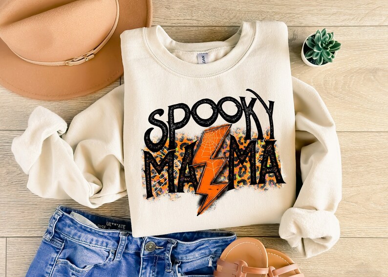 May include: Cream-colored sweatshirt with the text "SPOOKY MAMA" in black, orange, and leopard print. An orange lightning bolt with a spiderweb design is in the center. The sweatshirt is paired with blue jeans, a tan hat, and sandals.