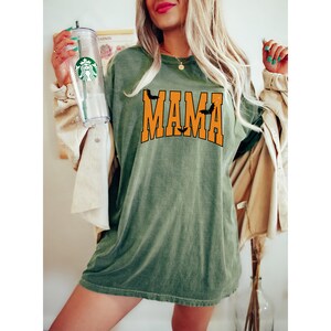 May include: Olive green t-shirt with the word "MAMA" in orange block letters, featuring bat silhouettes. The shirt is worn with a tan jacket and a clear tumbler with a green logo.