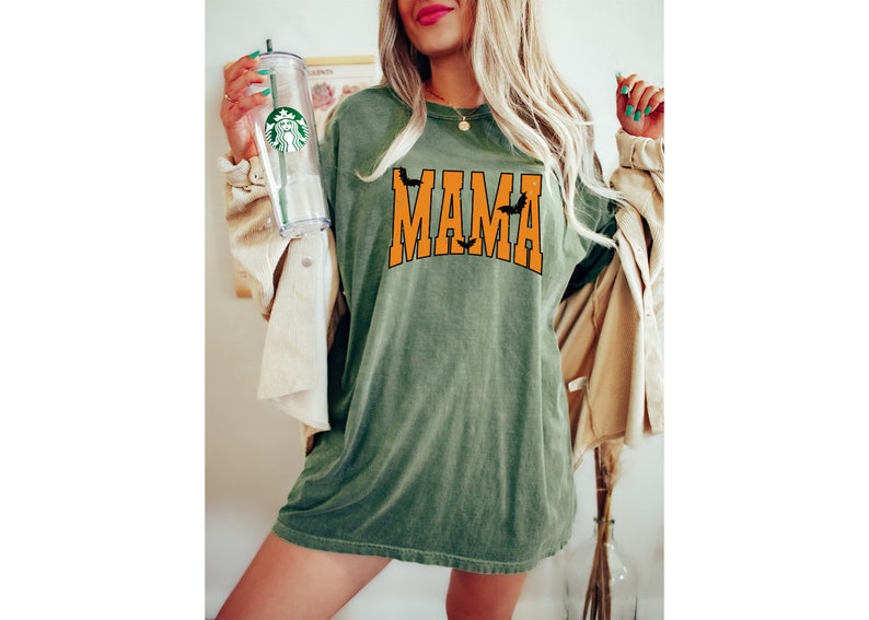 May include: Olive green t-shirt with the word "MAMA" in orange block letters, featuring bat silhouettes. The shirt is worn with a tan jacket and a clear tumbler with a green logo.