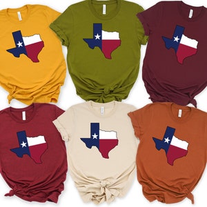 Op de afbeelding: Zes T-shirts in verschillende kleuren met een omtrek van de staat Texas in de kleuren van de vlag van Texas. De shirts zijn allemaal gevouwen en op een witte achtergrond gelegd.