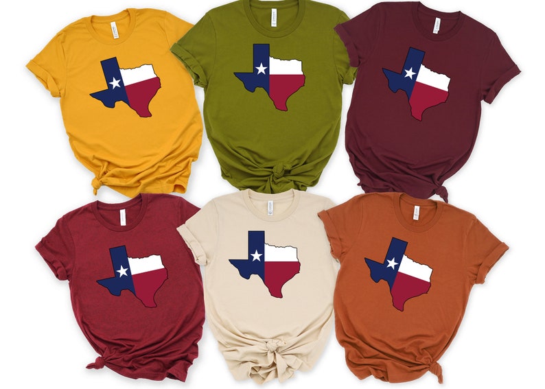 Op de afbeelding: Zes T-shirts in verschillende kleuren met een omtrek van de staat Texas in de kleuren van de vlag van Texas. De shirts zijn allemaal gevouwen en op een witte achtergrond gelegd.