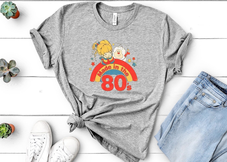 Puede incluir: Camiseta gris con un gr&aacute;fico retro de una ni&ntilde;a y un oso en un arco&iacute;ris con el texto "Made in the 80's".