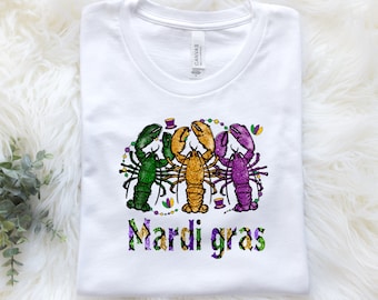 Op de afbeelding: Wit T-shirt met een afbeelding van drie kreeften die Mardi Gras-parels en hoeden dragen. De tekst "Mardi Gras" staat onder de kreeften.