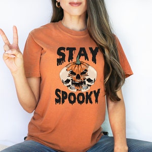 以下が含まれることがあります： ハロウィーンをテーマにしたオレンジ色のTシャツ。デザインは、2つのドクロと黒い文字で「STAY SPOOKY」と書かれたカボチャが特徴です。このTシャツは半袖で、クルーネックです。