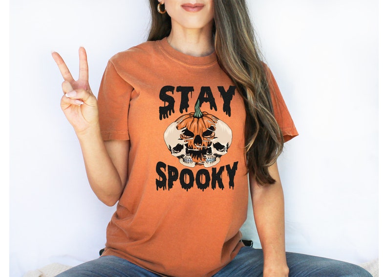 以下が含まれることがあります： ハロウィーンをテーマにしたオレンジ色のTシャツ。デザインは、2つのドクロと黒い文字で「STAY SPOOKY」と書かれたカボチャが特徴です。このTシャツは半袖で、クルーネックです。