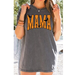 May include: Gray t-shirt with the word "MAMA" in orange block letters, arched across the chest. Black bat silhouettes are above and between the letters. A casual, Halloween-themed top.