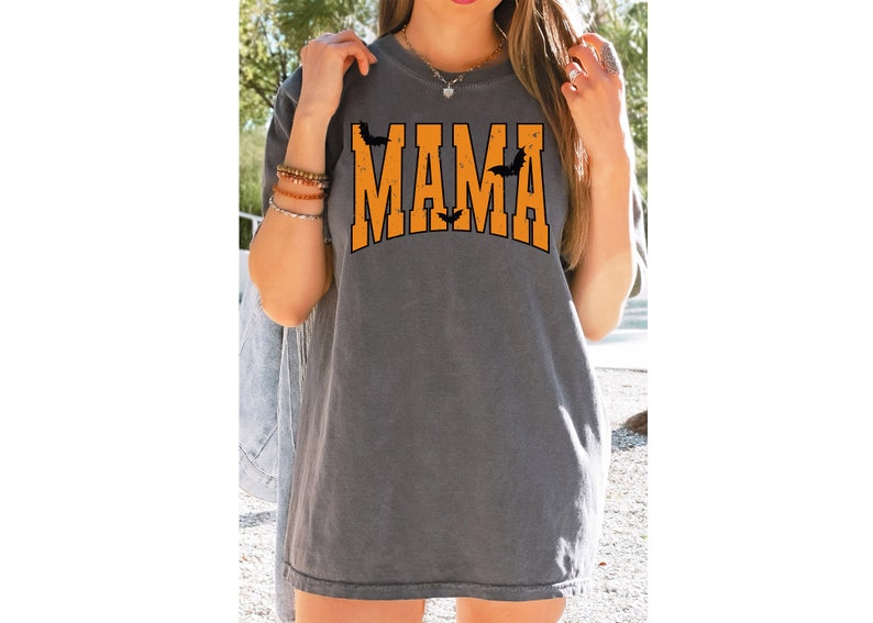 May include: Gray t-shirt with the word "MAMA" in orange block letters, arched across the chest. Black bat silhouettes are above and between the letters. A casual, Halloween-themed top.