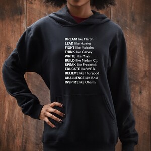 May include: Black hooded sweatshirt with white text that reads "DREAM like Martin, LEAD like Harriet, FIGHT like Malcolm, THINK like Garvey, WRITE like Maya, BUILD like Madam C.J., SPEAK like Frederick, EDUCATE like W.E.B., BELIEVE like Thurgood, CHALLENGE like Rosa, INSPIRE like Obama."