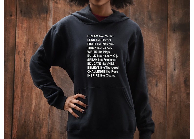 May include: Black hooded sweatshirt with white text that reads "DREAM like Martin, LEAD like Harriet, FIGHT like Malcolm, THINK like Garvey, WRITE like Maya, BUILD like Madam C.J., SPEAK like Frederick, EDUCATE like W.E.B., BELIEVE like Thurgood, CHALLENGE like Rosa, INSPIRE like Obama."