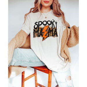 May include: Cream-colored t-shirt with the text "Spooky Mama" in black and leopard print, featuring an orange lightning bolt. The shirt is part of a Halloween-themed collection.