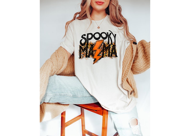 May include: Cream-colored t-shirt with the text "Spooky Mama" in black and leopard print, featuring an orange lightning bolt. The shirt is part of a Halloween-themed collection.