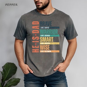 May include: A gray t-shirt with a vintage-style graphic that reads "He is Dad" and lists qualities like "Brave", "Warrior", "Smart", and "Wise" with biblical references.