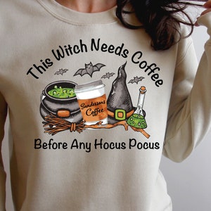 May include: Beige sweatshirt with a Halloween-themed graphic. The design includes a witch's cauldron, coffee cup, witch hat, and potion bottle. Text reads "This Witch Needs Coffee Before Any Hocus Pocus."