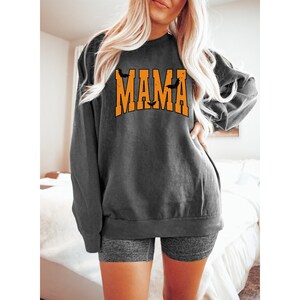 May include: Dark gray sweatshirt with the word "MAMA" in orange block letters, with bat silhouettes. The sweatshirt has long sleeves and a crew neck. The overall style is casual and relaxed.