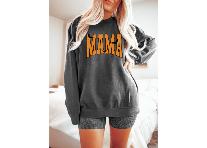 May include: Dark gray sweatshirt with the word "MAMA" in orange block letters, with bat silhouettes. The sweatshirt has long sleeves and a crew neck. The overall style is casual and relaxed.
