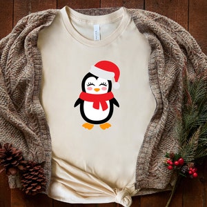 May include: A beige t-shirt with a cute cartoon penguin wearing a red Santa hat and scarf. The penguin has black eyes, a red nose, and a white body.