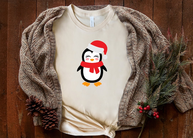 May include: A beige t-shirt with a cute cartoon penguin wearing a red Santa hat and scarf. The penguin has black eyes, a red nose, and a white body.