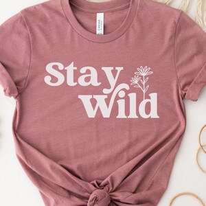 May include: A mauve t-shirt with the text "Stay Wild" in white lettering with a stylized flower graphic.