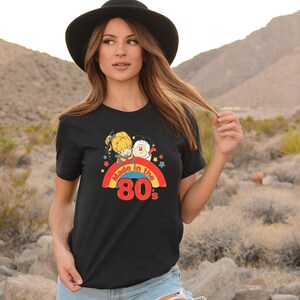 Puede incluir: Camiseta negra con un gr&aacute;fico de arco&iacute;ris y estrellas. El gr&aacute;fico presenta dos personajes de dibujos animados y el texto "Made in the 80's".