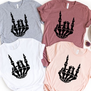 Puede incluir: Cuatro camisetas en gris, blanco, malva y rosa claro, cada una con una mano de esqueleto negra haciendo un gesto de rock and roll. Las camisetas son de manga corta y tienen cuello redondo.