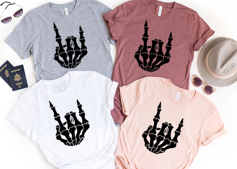 Puede incluir: Cuatro camisetas en gris, blanco, malva y rosa claro, cada una con una mano de esqueleto negra haciendo un gesto de rock and roll. Las camisetas son de manga corta y tienen cuello redondo.