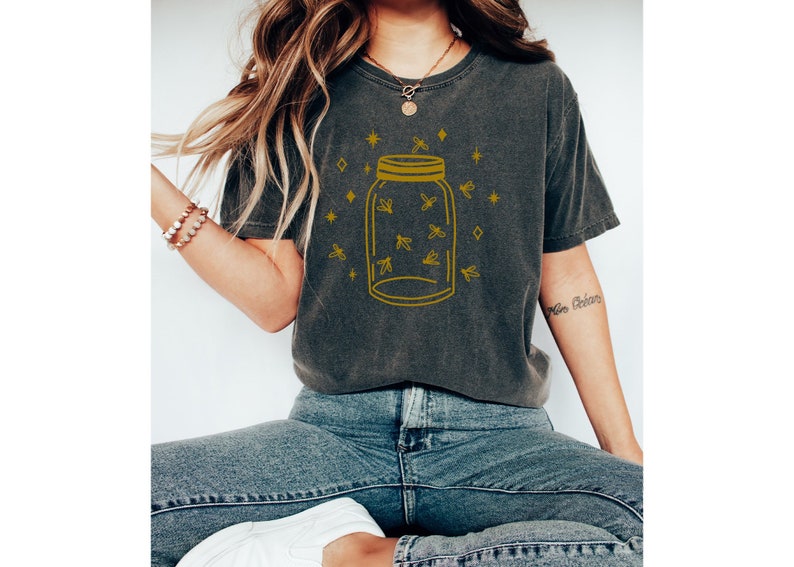 以下が含まれることがあります： 暗いグレーのTシャツに、金色の輪郭で描かれた、ホタルでいっぱいのメイソンジャーのデザイン。デザインは、金色の星とダイヤモンドで囲まれています。
