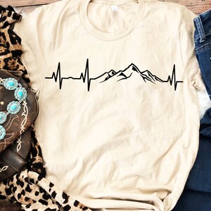 K&ouml;nnte beinhalten: Ein beiges T-Shirt mit einem schwarzen Bergketten-Motiv und einer Herzschlaglinie. Das Motiv ist mittig auf dem Shirt platziert.