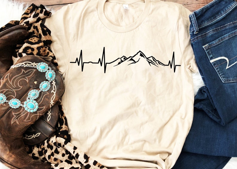 K&ouml;nnte beinhalten: Ein beiges T-Shirt mit einem schwarzen Bergketten-Motiv und einer Herzschlaglinie. Das Motiv ist mittig auf dem Shirt platziert.