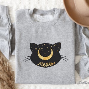 May include: A heather grey sweatshirt with a black cat silhouette design. The cat has a crescent moon with gold stars on its head and the word "meow" underneath.
