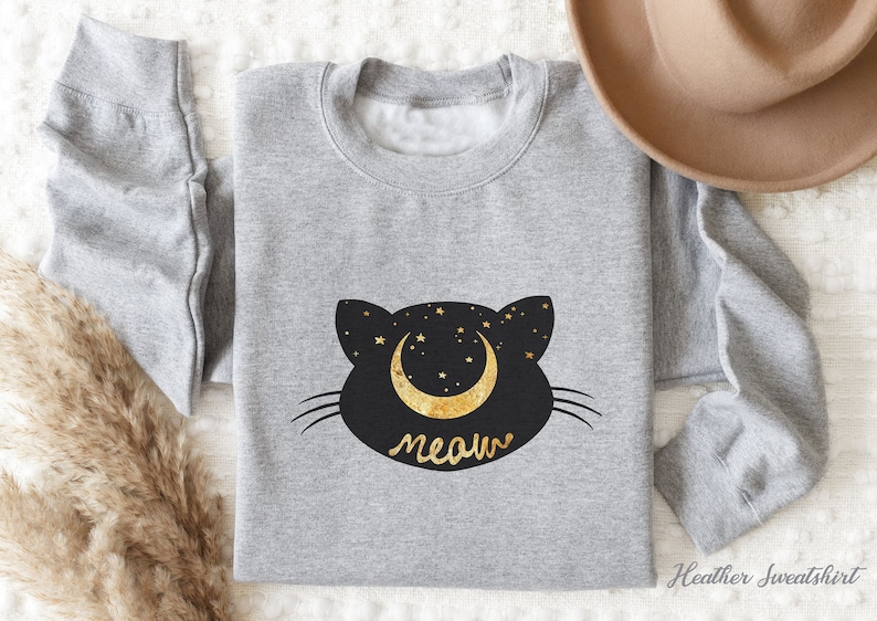 May include: A heather grey sweatshirt with a black cat silhouette design. The cat has a crescent moon with gold stars on its head and the word "meow" underneath.