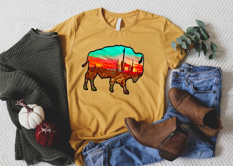 May include: A mustard yellow t-shirt with a black silhouette of a bison. The bison is filled with a colorful sunset scene with cacti and a blue sky.