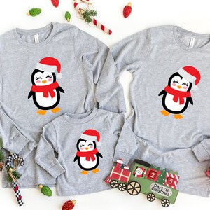 May include: A set of three gray long-sleeved shirts with a cute penguin wearing a red Santa hat and scarf design. The shirts are perfect for a family Christmas outfit.