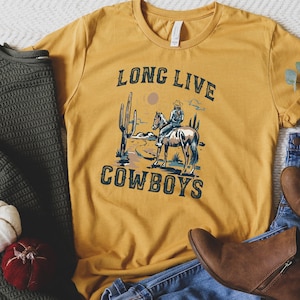May include: A mustard yellow t-shirt with a graphic of a cowboy riding a horse in a desert landscape. The text "Long Live Cowboys" is printed on the shirt.