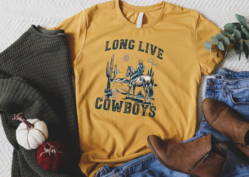 May include: A mustard yellow t-shirt with a graphic of a cowboy riding a horse in a desert landscape. The text "Long Live Cowboys" is printed on the shirt.