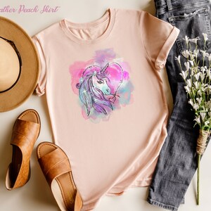 May include: A heather peach t-shirt with a watercolor unicorn design in shades of pink, purple, and blue. The unicorn is inside a heart shape.