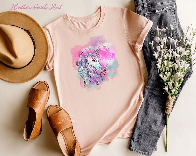 May include: A heather peach t-shirt with a watercolor unicorn design in shades of pink, purple, and blue. The unicorn is inside a heart shape.