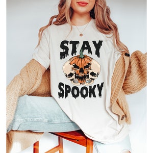 以下が含まれることがあります： ハロウィーンをテーマにしたグラフィックのクリーム色のTシャツ。デザインは、2つのスカルと「STAY SPOOKY」という言葉が黒色の滴るフォントで描かれたカボチャが特徴です。このシャツはカジュアルな半袖スタイルです。