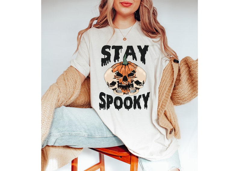 以下が含まれることがあります： ハロウィーンをテーマにしたグラフィックのクリーム色のTシャツ。デザインは、2つのスカルと「STAY SPOOKY」という言葉が黒色の滴るフォントで描かれたカボチャが特徴です。このシャツはカジュアルな半袖スタイルです。