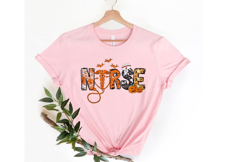 May include: A light pink t-shirt with the word "NURSE" in a Halloween-themed design. The letters are filled with pumpkins, bats, and a nurse's cap. A medical caduceus and stethoscope are also featured.