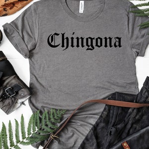 May include: A grey t-shirt with the word "Chingona" printed in black gothic lettering. The shirt is laid out on a white surface with a pair of black boots, a brown leather strap, and a pair of black jeans.