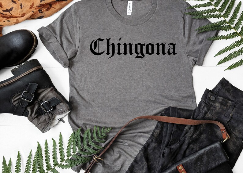May include: A grey t-shirt with the word "Chingona" printed in black gothic lettering. The shirt is laid out on a white surface with a pair of black boots, a brown leather strap, and a pair of black jeans.
