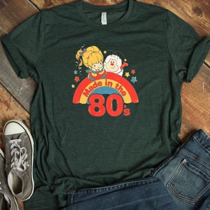 Puede incluir: Una camiseta verde oscuro con un arco&iacute;ris y el texto "Made in the 80's". El arco&iacute;ris tiene una ni&ntilde;a de dibujos animados y una criatura blanca en la parte superior.