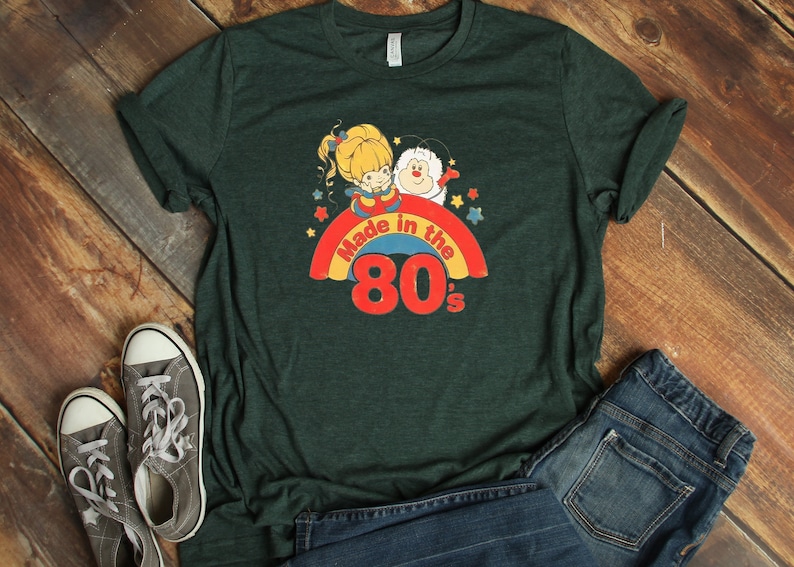 Puede incluir: Una camiseta verde oscuro con un arco&iacute;ris y el texto "Made in the 80's". El arco&iacute;ris tiene una ni&ntilde;a de dibujos animados y una criatura blanca en la parte superior.