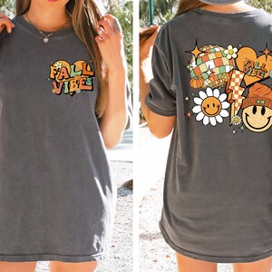 May include: A gray oversized t-shirt with a graphic print on the front that says "Fall Vibes" in a heart shape and a graphic print on the back that includes a smiley face flower, a disco ball, a pumpkin, a lightning bolt, and a smiley face wearing a beanie.