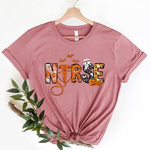 May include: Mauve t-shirt with the word "NURSE" in a Halloween-themed design. The letters are decorated with pumpkins, bats, and a medical caduceus. The shirt is displayed on a wooden hanger.